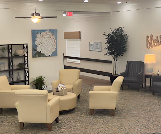 A cozy living room area in a senior living facility with four beige armchairs arranged around a round ottoman. The room features a ceiling fan with a light, a large floral painting on the wall, a tall black shelving unit, a potted plant, and a gray armchair near a side table with a lamp. The walls are light-colored, and there is a carpeted floor.