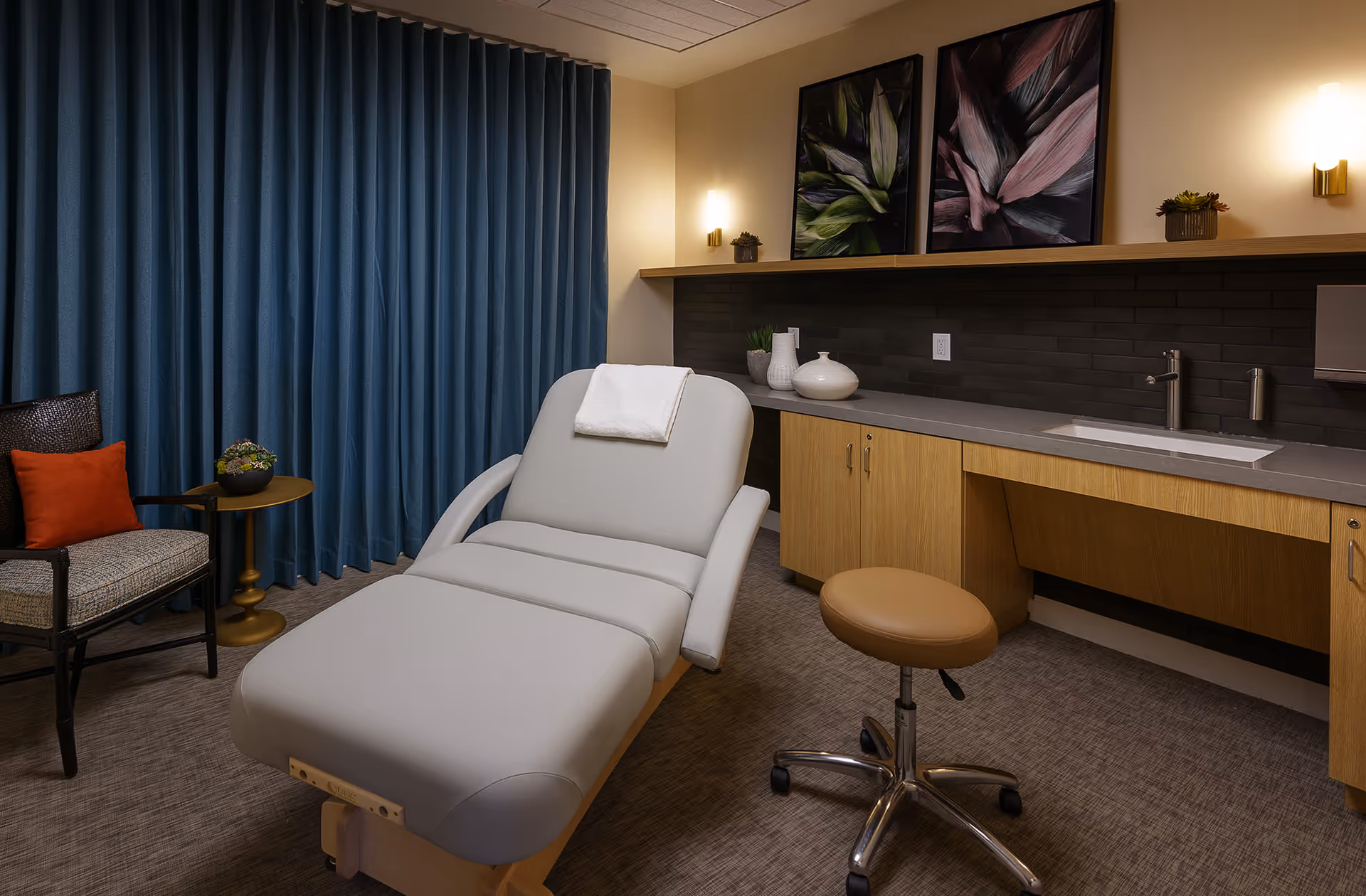 A modern treatment room featuring a padded reclining chair with a white towel draped over the headrest, a tan rolling stool, a wooden cabinet with a gray countertop and built-in sink, decorative vases, two framed botanical prints on the wall, a dark tiled backsplash, a blue curtain, and a chair with an orange cushion next to a small round table with a plant.