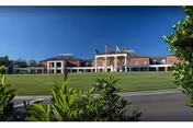 Front exterior view of a large brick building with white columns and multiple flags flying on poles in front. The building is set behind a well-maintained grassy area with shrubs and trees under a clear blue sky.