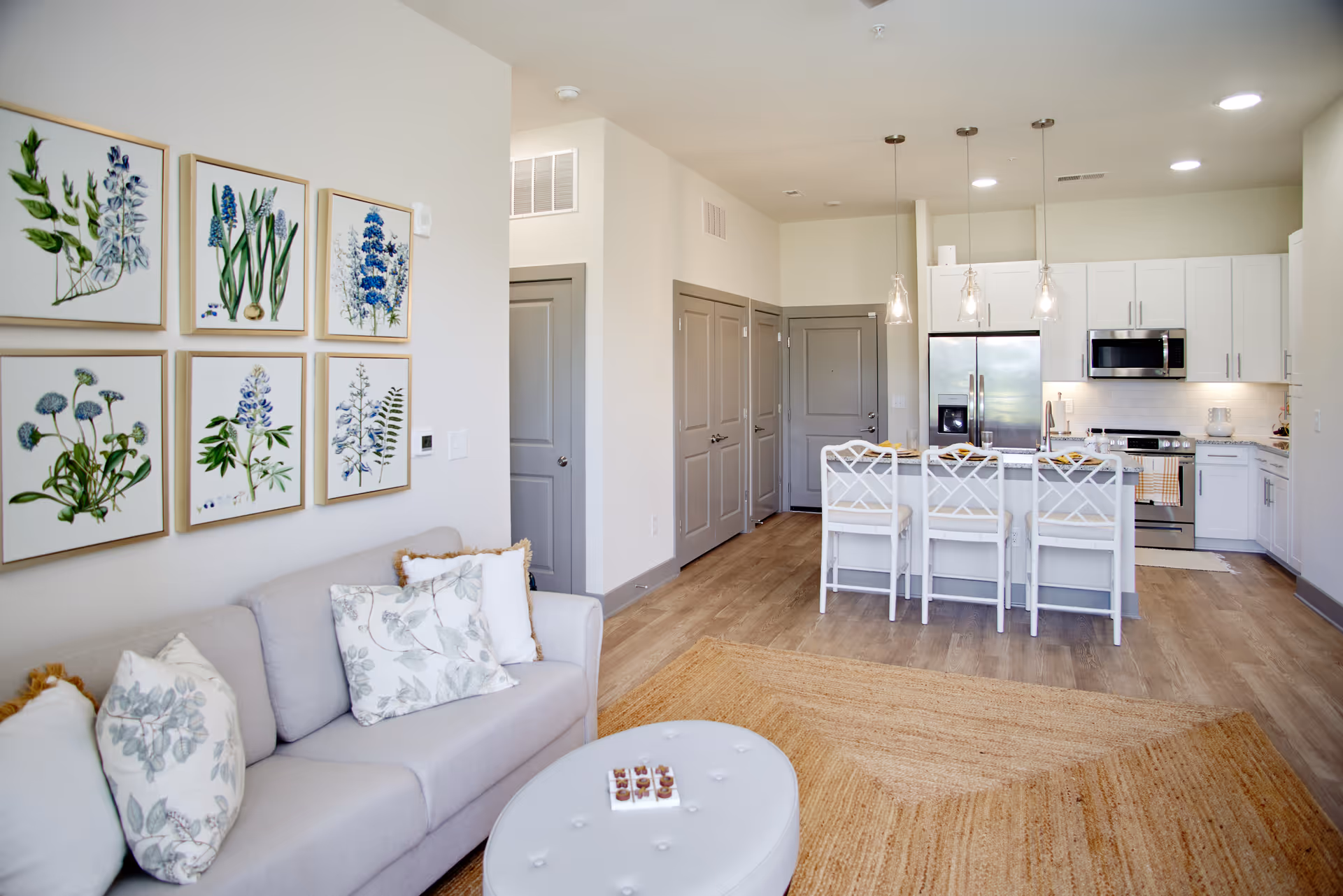 A modern open-concept living room and kitchen area with a light gray sofa adorned with floral pillows, a round white tufted ottoman, and a natural fiber rug. The kitchen features white cabinetry, stainless steel appliances, a granite countertop island with three white bar stools, and pendant lighting. Botanical artwork is displayed on the wall above the sofa.