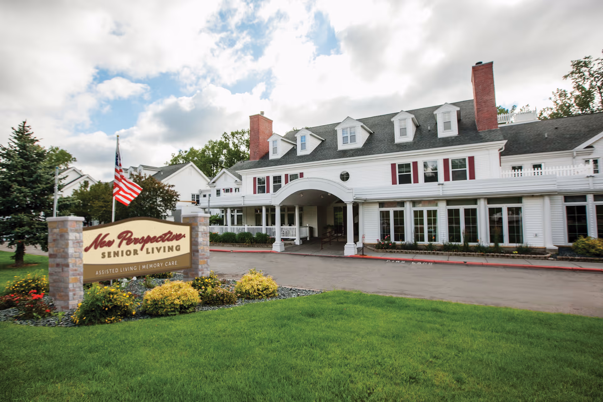 Exterior view of New Perspective Senior Living facility in Roseville, showing a large white building with multiple windows, two brick chimneys, and a covered entrance. In front of the building is a landscaped area with green grass, bushes, flowers, and a sign that reads 'New Perspective Senior Living Assisted Living | Memory Care'. An American flag is also visible near the sign.