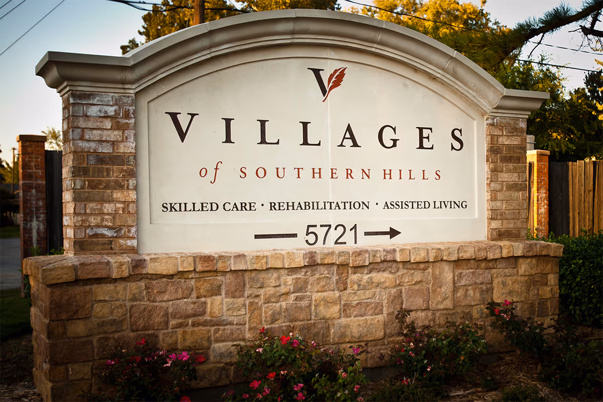 Stone and brick sign for The Villages of Southern Hills, indicating skilled care, rehabilitation, and assisted living services, with the number 5721 and an arrow pointing right, surrounded by greenery and flowers.