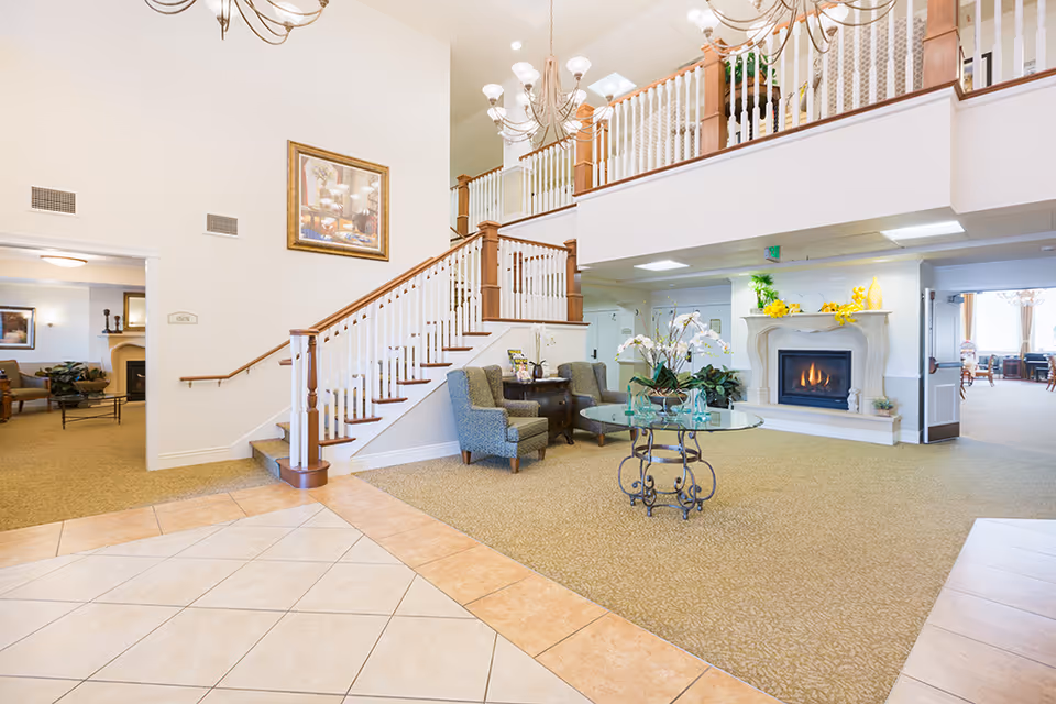 Bright and spacious senior living facility interior with a staircase featuring wooden handrails and white balusters. There are two upholstered chairs and a small table with flowers near the staircase. A glass-top round table with a floral arrangement sits on a carpeted floor. A fireplace with a mantel decorated with yellow flowers and greenery is visible. The area is well-lit with chandeliers and recessed lighting, and there is an open doorway leading to another room with more seating and a fireplace.