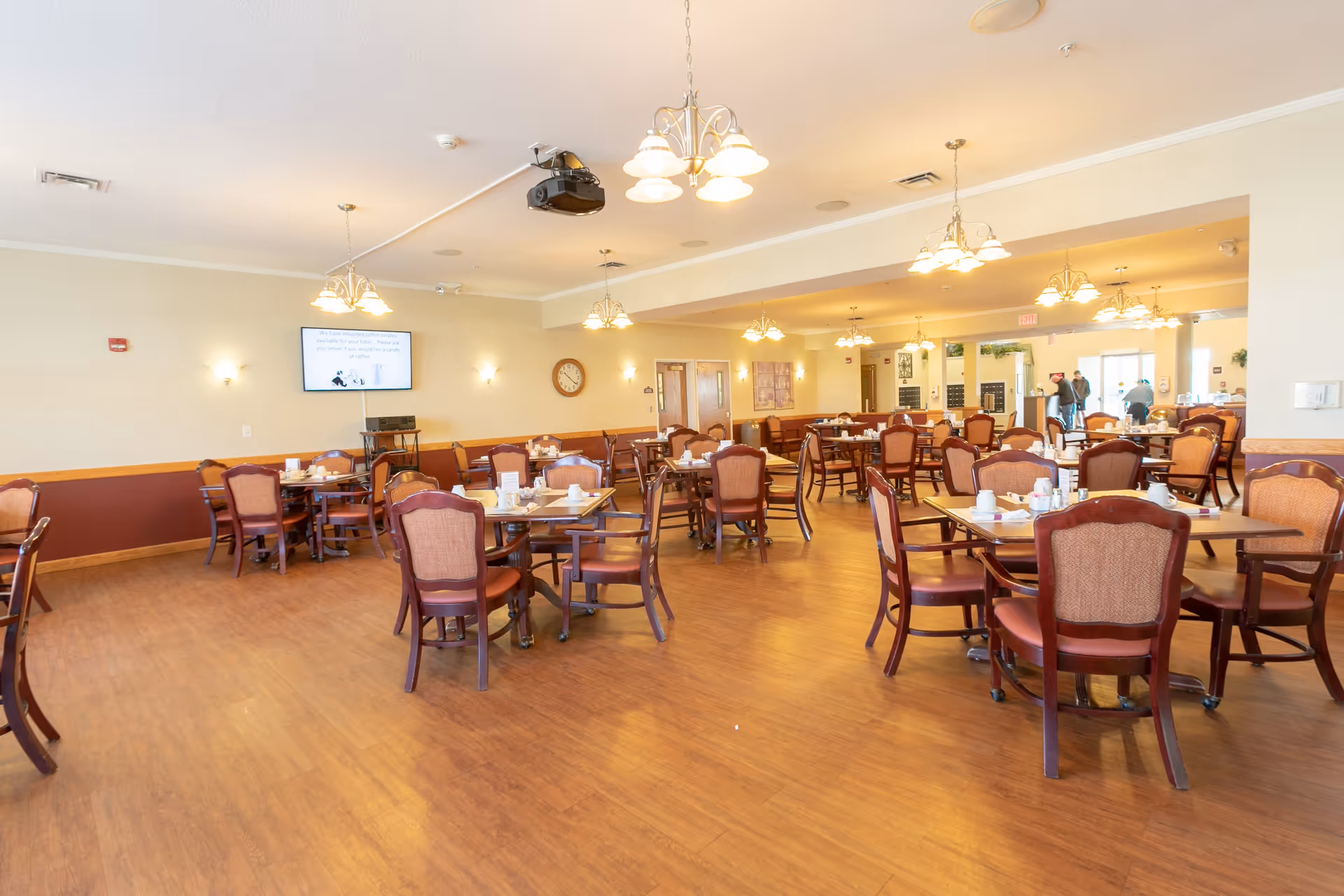 A spacious dining room with multiple wooden tables and chairs arranged neatly. The tables have cups, plates, and napkins set on them. The room has warm lighting from ceiling chandeliers and wall sconces. A clock and a television screen are mounted on the wall. The floor is wooden, and there are a few people visible near the entrance in the background.