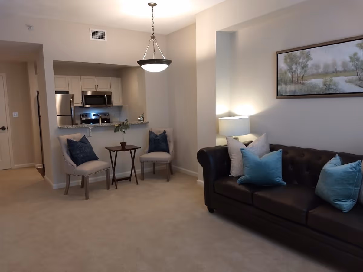 A cozy living area featuring a dark brown leather sofa with blue and white throw pillows, a side table with a lamp, two beige chairs with dark blue pillows, and a small table with a potted plant. In the background, there is a kitchen with stainless steel appliances including a refrigerator, microwave, and stove, and white cabinetry. The walls are painted light beige, and a landscape painting hangs above the sofa.