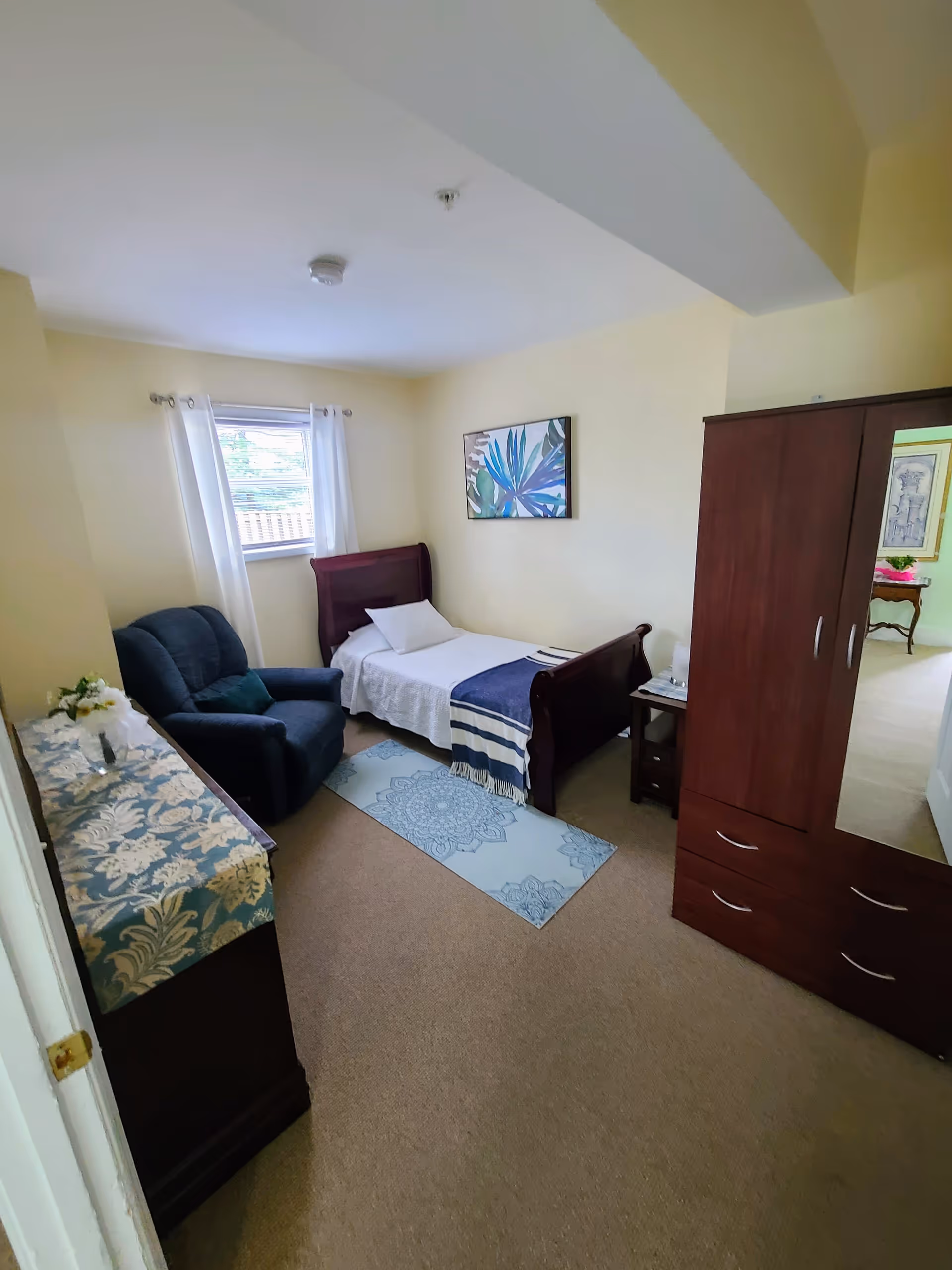 A small bedroom with beige walls and carpet. The room contains a single wooden bed with white bedding and a blue striped throw blanket, a blue upholstered armchair, a wooden dresser with a floral runner and a small flower arrangement, and a wooden wardrobe with a mirrored door. A window with white curtains lets in natural light, and a framed artwork with blue and green leaves hangs on the wall above the bed.