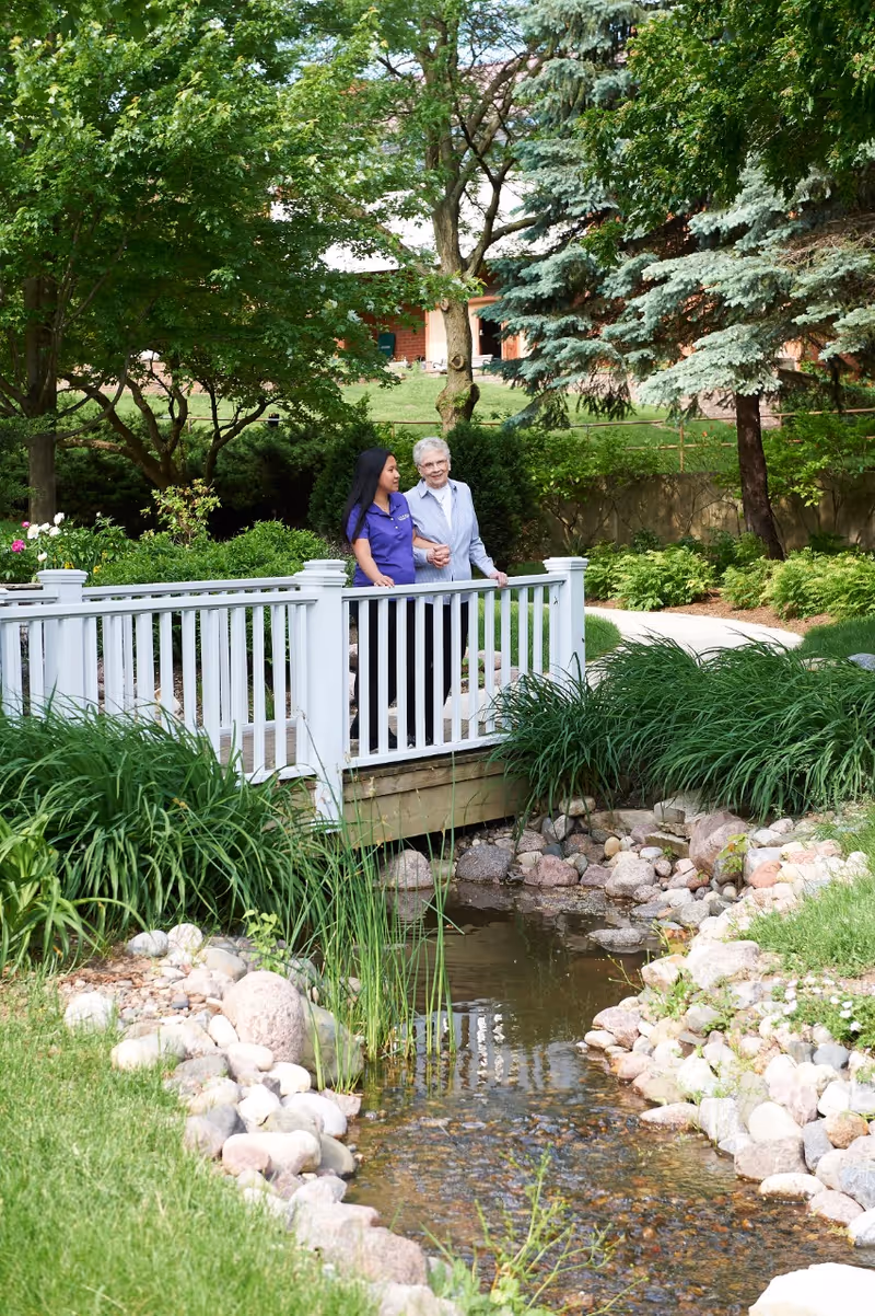 An older woman and a caregiver standing on a white wooden bridge over a small stream in a landscaped garden.