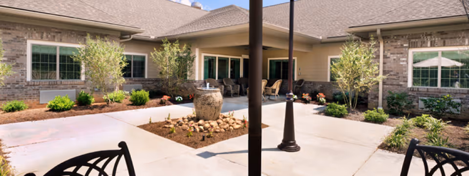 Outdoor patio area at Bedford Care Center Of Picayune featuring a central decorative water fountain surrounded by landscaping with small bushes and trees. The patio is paved with concrete and has several chairs arranged under a covered area attached to the building with brick and siding exterior walls and multiple windows.