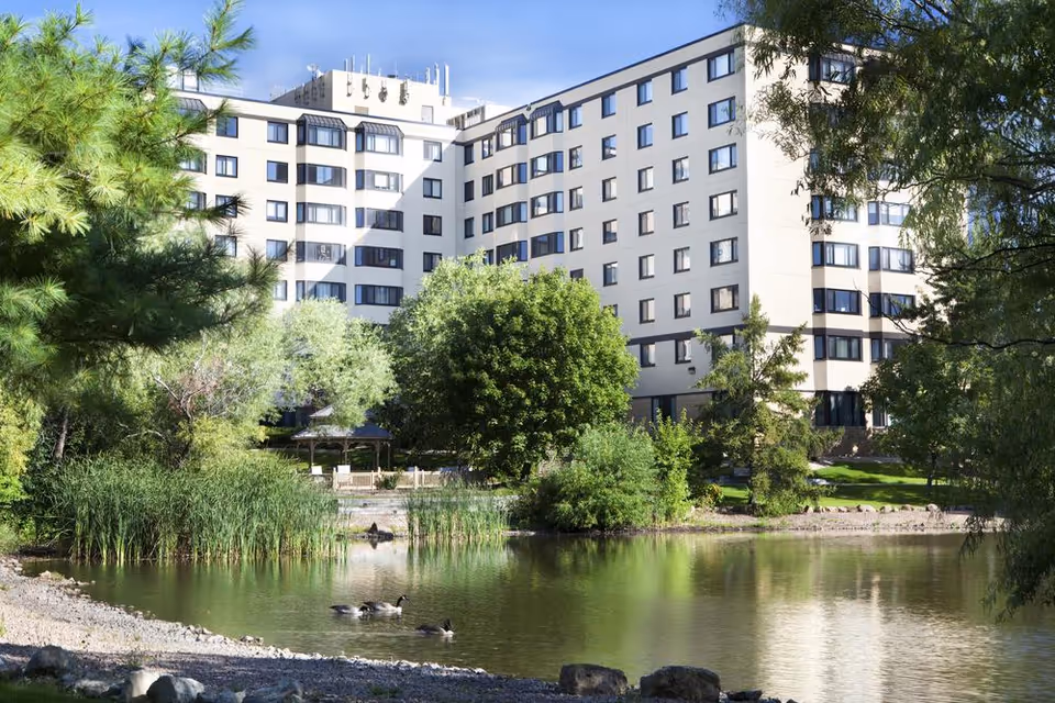A multi-story senior living facility building named Knollwood Place Apartments is visible behind a serene pond with ducks swimming. The foreground features lush greenery, trees, and a small gazebo near the water's edge.