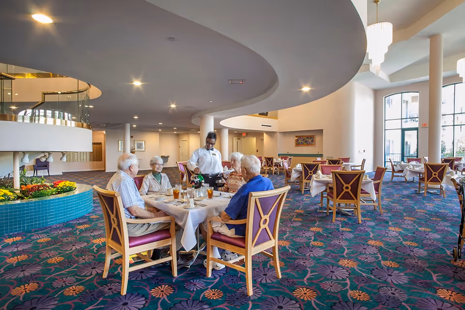 A server attends to a group of seniors seated at a table in a spacious, well-lit dining room with a patterned carpet.