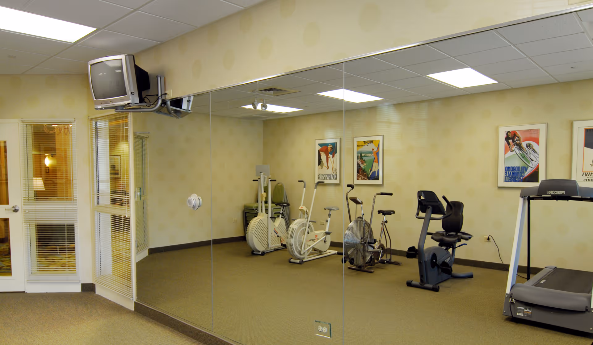 Small fitness room with exercise bikes, a treadmill, a mirrored wall, and a wall-mounted TV.