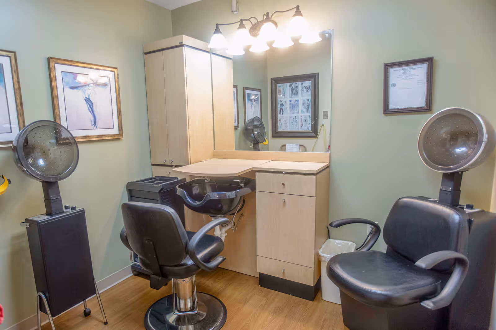 Small salon-style hair station with two styling chairs, hooded dryers, a shampoo bowl, mirror, and cabinets.