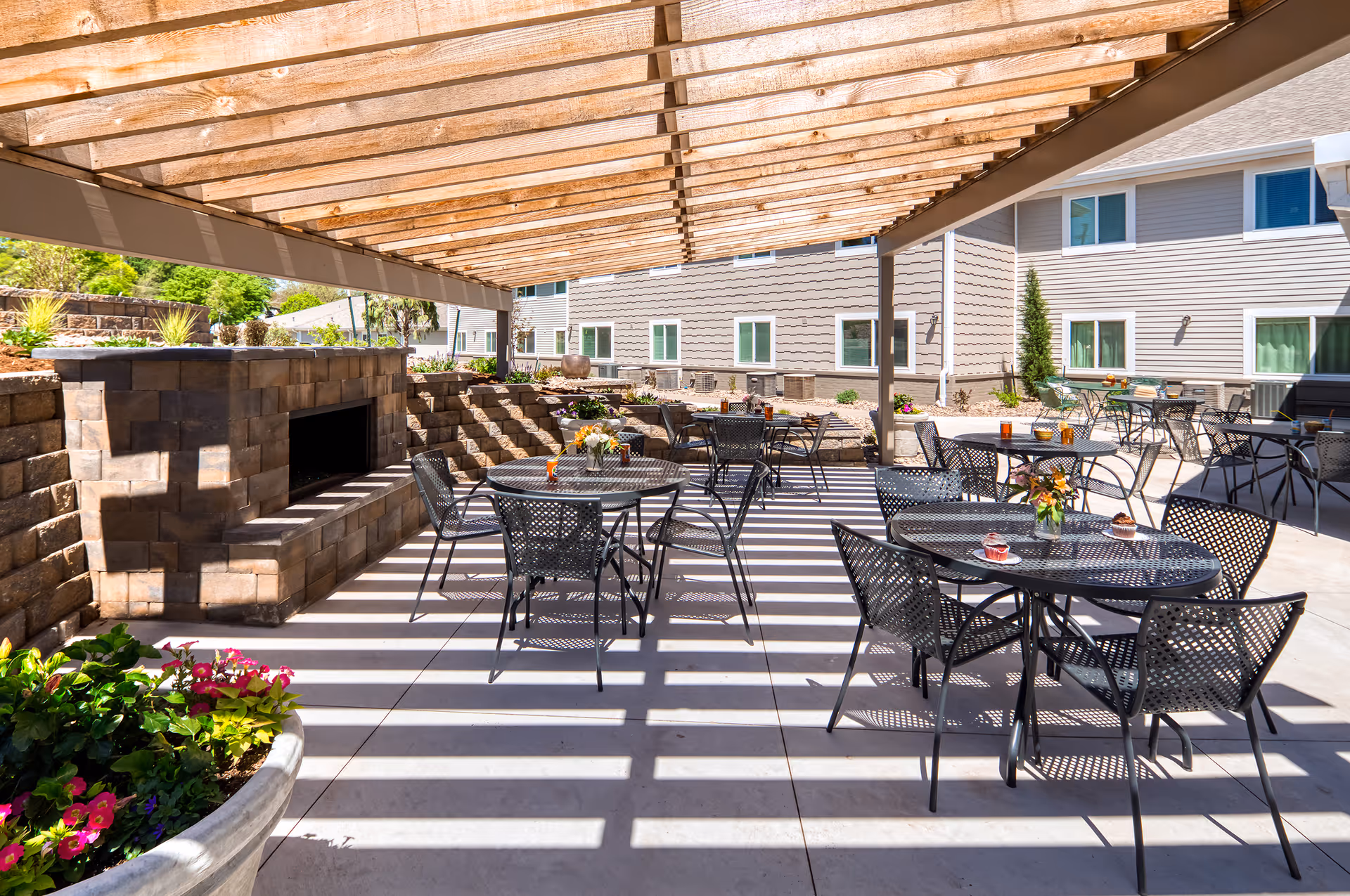 Outdoor covered patio at Parkwood Village with a wooden pergola, metal dining tables and chairs, potted plants, and a stone fireplace in front of the building.