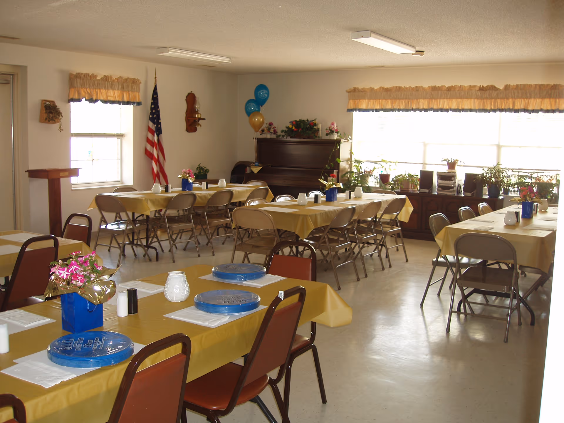 Community dining room with folding tables and chairs covered in yellow tablecloths, place settings, an American flag, and a piano by the windows.