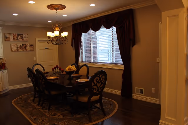 A warmly lit dining room with a dark wooden dining table set for six. The table has a centerpiece with bananas and candles. The room features a chandelier, a large window with dark curtains, a patterned rug under the table, and framed artwork on the wall.