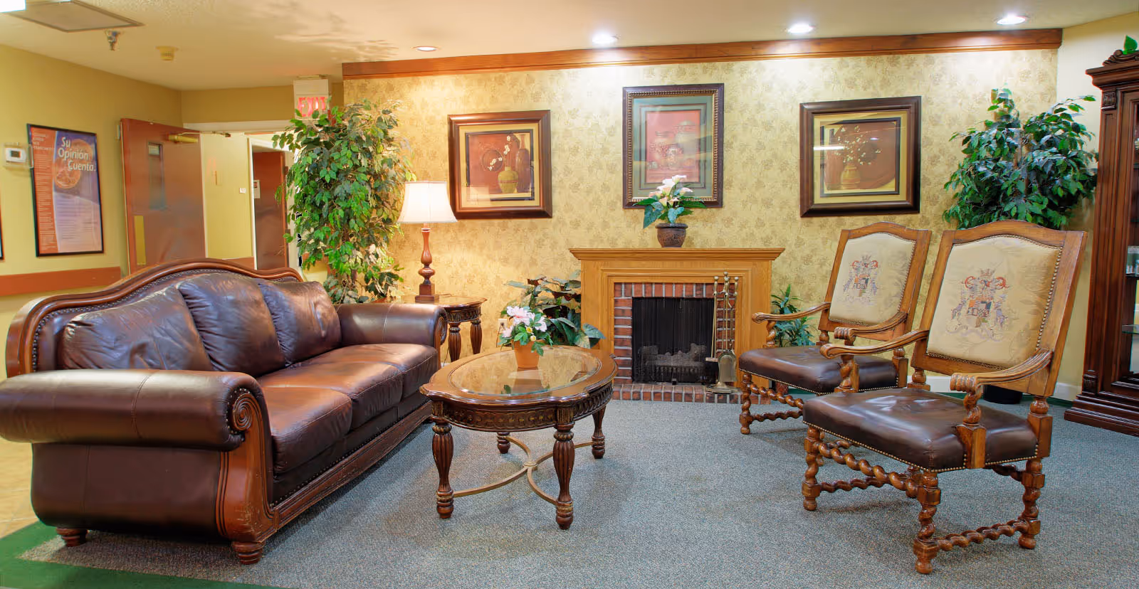 A cozy living room area with a brown leather sofa, two wooden armchairs with upholstered backs and leather seats, a wooden coffee table with a glass top, a side table with a lamp, and a fireplace with decorative plants and framed artwork on the wall.