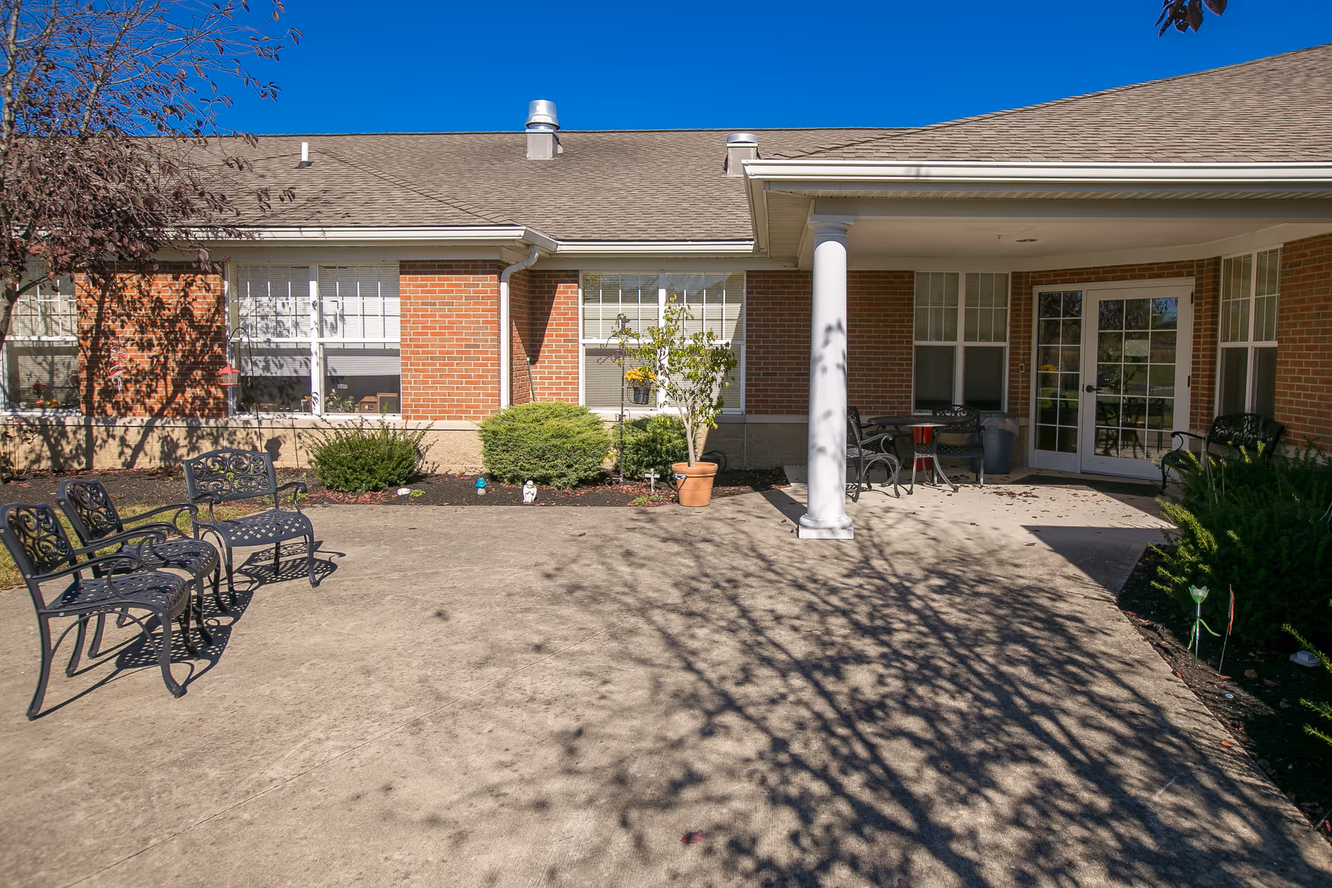 Outdoor patio area of a brick building with a concrete floor, featuring black metal chairs and benches, potted plants, and a white column supporting a roof overhang. The sky is clear and blue, and shadows of tree branches are cast on the ground.