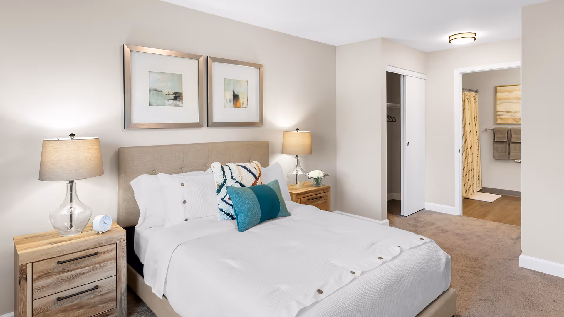 A neatly made bed with white bedding and decorative pillows in a bedroom. There are two wooden nightstands on either side of the bed, each with a lamp. Two framed abstract paintings hang above the bed. An open closet with sliding doors and an entrance to a bathroom with towels and a shower curtain are visible in the background.