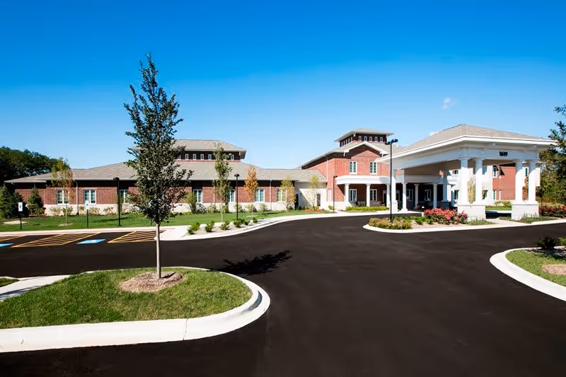 Exterior view of HarborChase of Naperville, showing a large brick building with a covered entrance, surrounded by a paved driveway, landscaped greenery, and small trees under a clear blue sky.