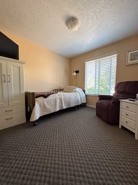 A small bedroom with a single bed covered with a white quilt, a brown recliner chair, a white nightstand with drawers, a window with white blinds letting in natural light, and a white wardrobe with a TV on top.