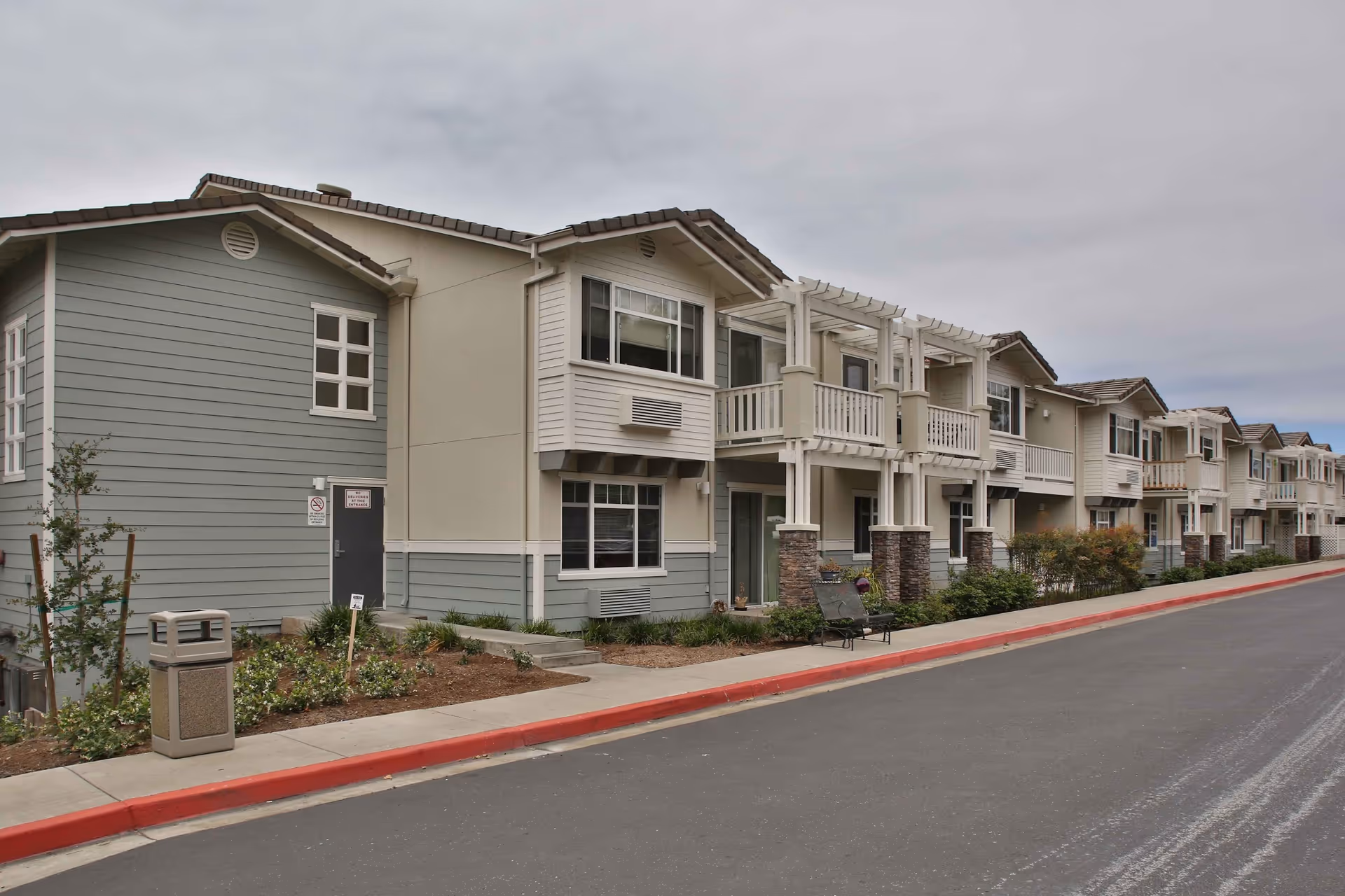 Exterior view of a multi-unit residential building with balconies, beige and light gray siding, and a paved road with a red curb in front. There are small landscaped areas with plants and a trash can near the sidewalk.
