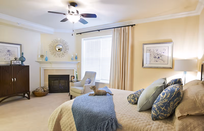 Sunlit, neatly staged bedroom with a made bed and decorative pillows, a fireplace, armchair, and ceiling fan.