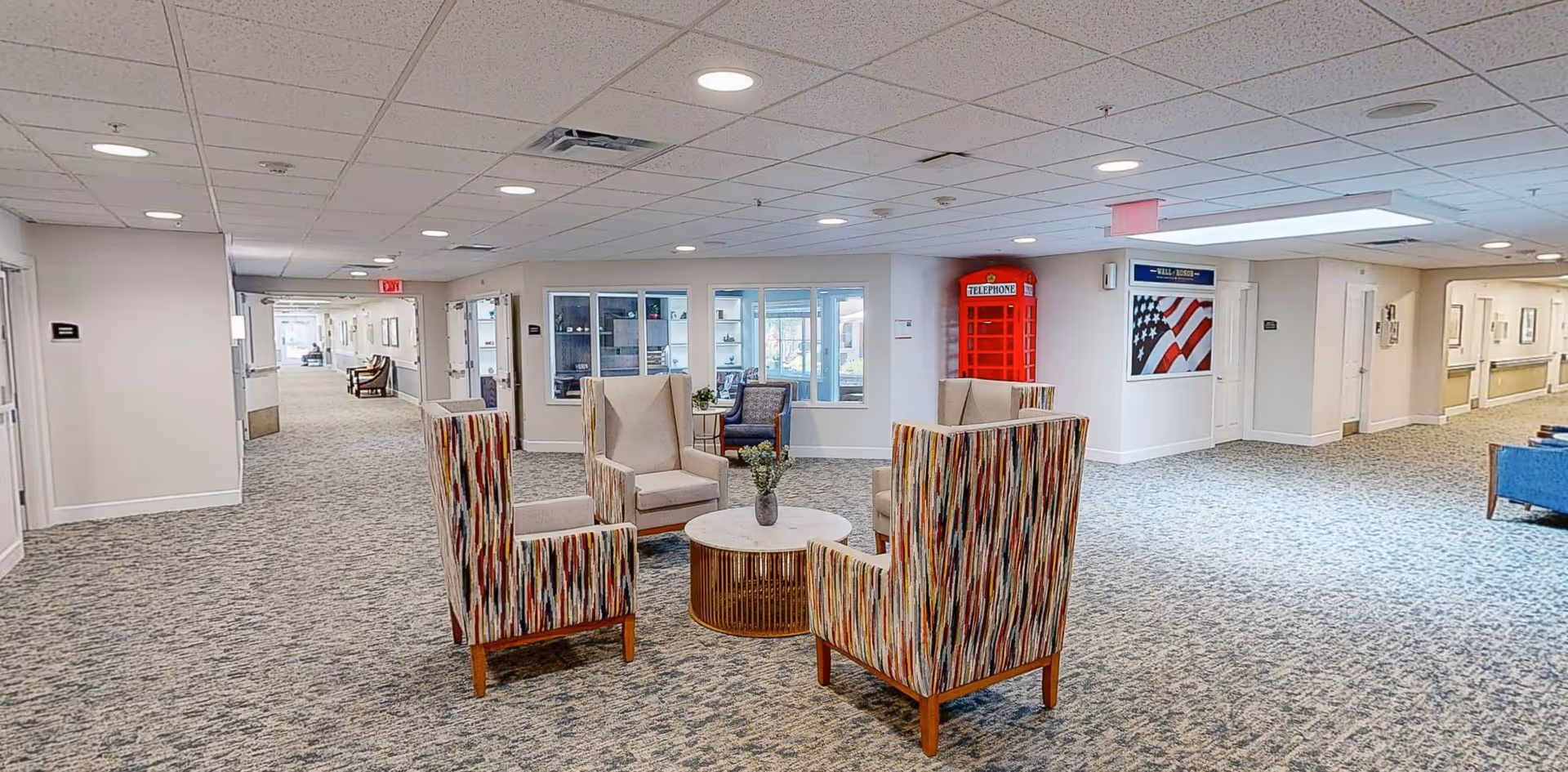 A spacious, well-lit common area in a senior living facility with patterned armchairs arranged around a round coffee table with a small plant. The area has carpeted floors, white walls, and a ceiling with recessed lighting. In the background, there is a red British-style telephone booth and a framed American flag on the wall. Hallways extend to the left and right with additional seating visible.