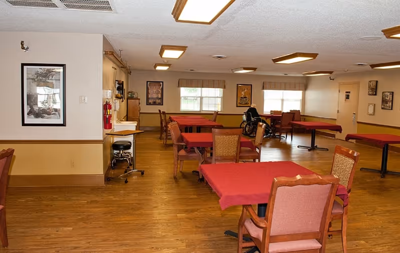 Communal dining room with multiple tables covered in red tablecloths, wooden chairs, and windows along the back wall.