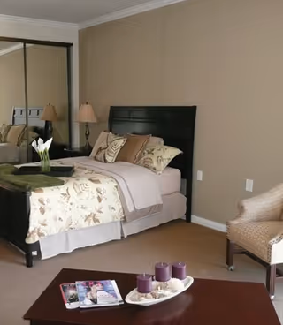 A neatly made bed with floral patterned bedding and multiple pillows in a bedroom. There is a wooden headboard, two bedside lamps, a mirrored closet door, a beige armchair, and a wooden coffee table with three purple candles and magazines on it.