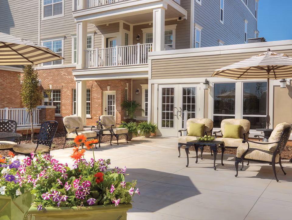 Outdoor patio area at a senior living facility with cushioned chairs, tables, umbrellas, and colorful flower planters. The building exterior features brick and siding with multiple windows and a balcony.