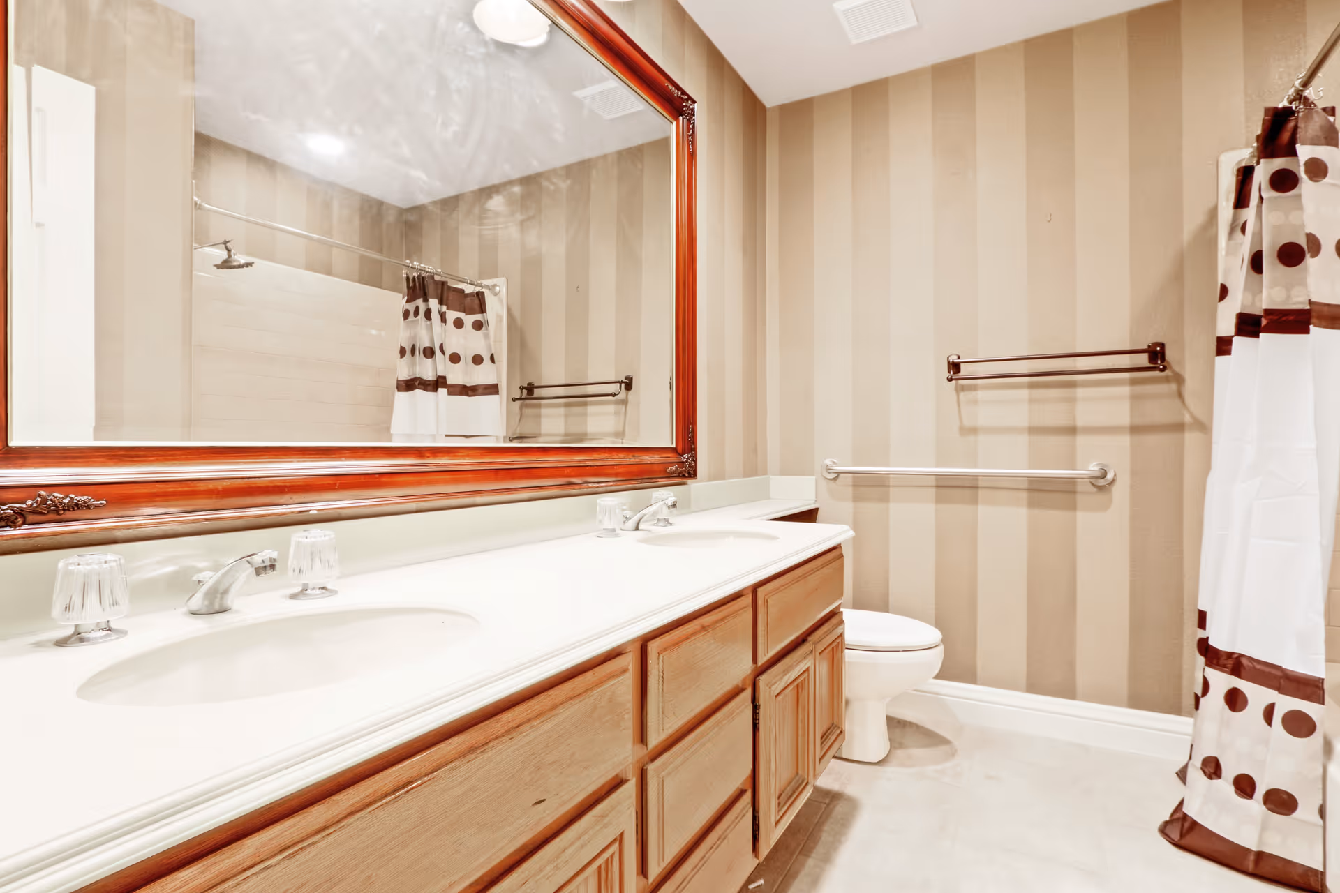 A bathroom with a double sink vanity featuring wooden cabinets and a large framed mirror above. The walls have beige vertical stripes. There is a toilet with a grab bar next to it and a shower with a curtain that has a brown and white polka dot pattern.