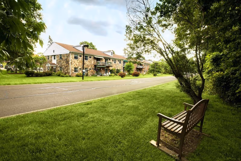A peaceful outdoor scene at Spring House Estates featuring a wooden bench on a grassy area next to a tree, with a paved road and a two-story stone building with balconies in the background under a partly cloudy sky.