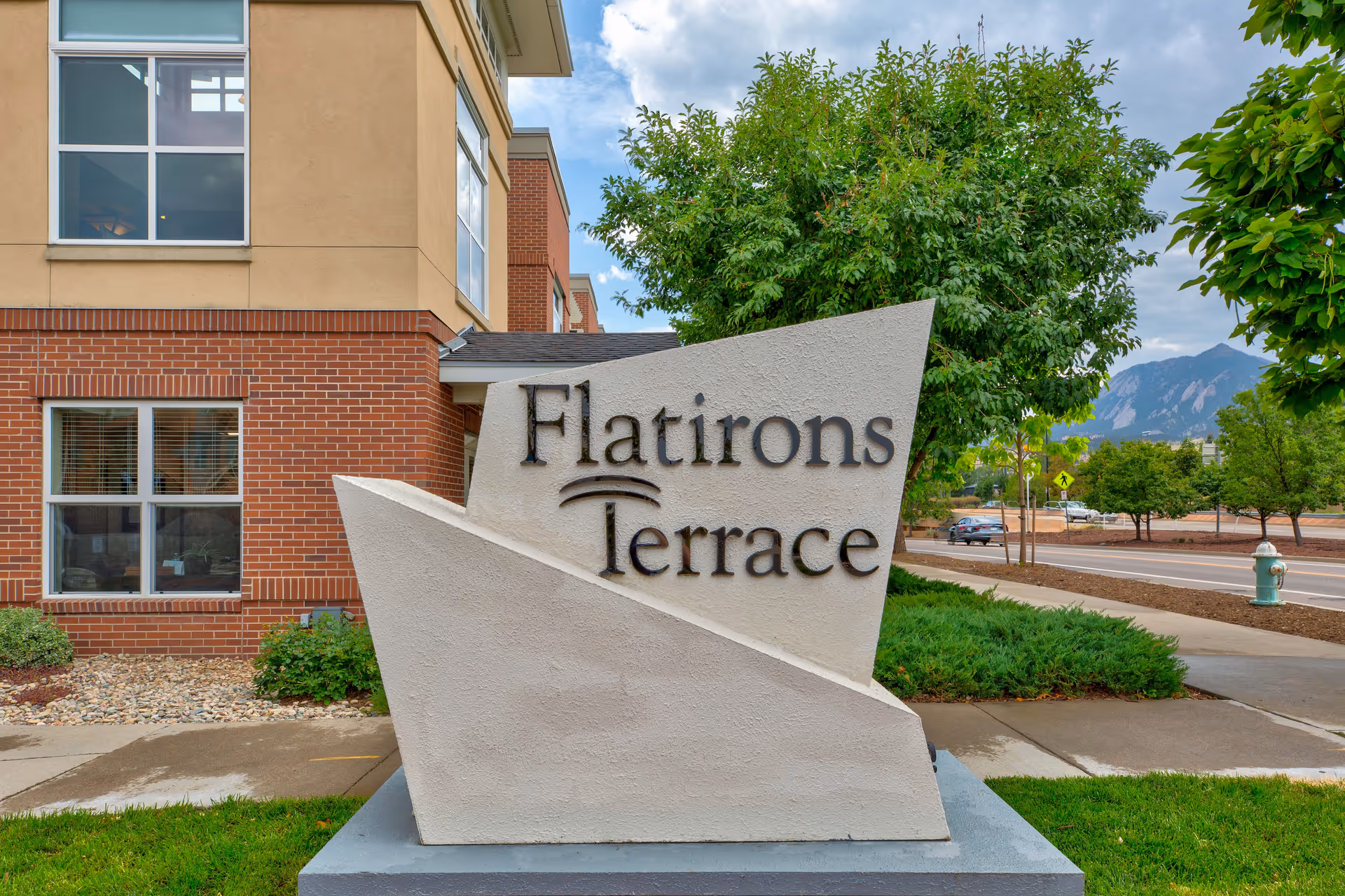 A sculpted stone sign that reads "Flatirons Terrace" stands on a lawn in front of a brick-and-stucco building with trees and distant mountains.