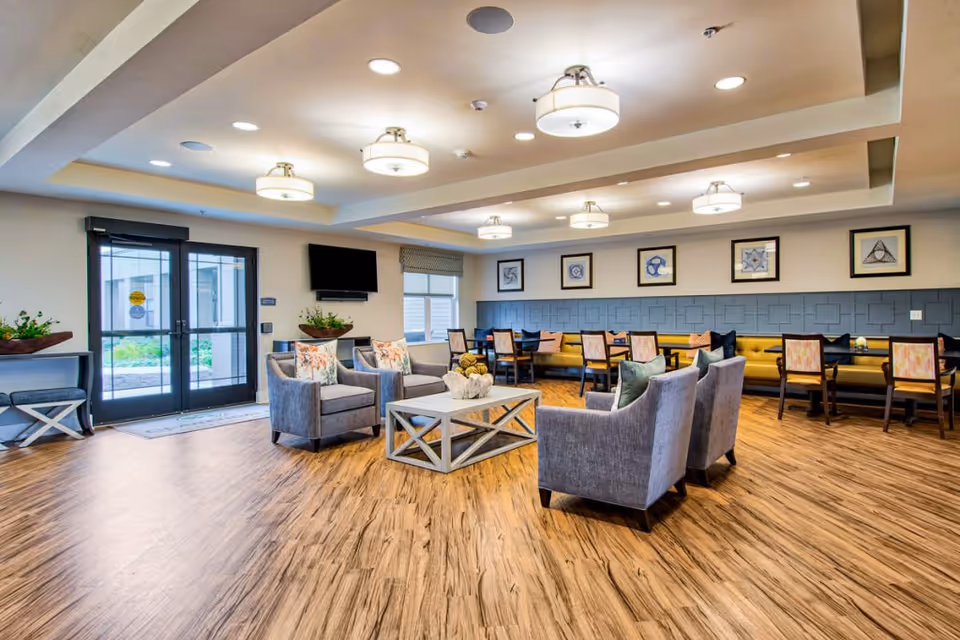 A spacious and well-lit common area with wooden flooring, featuring a seating arrangement of four gray armchairs around a white coffee table in the center. Along the back wall, there is a long yellow cushioned bench with multiple dining tables and chairs. The walls are decorated with framed geometric artwork, and ceiling lights provide ample illumination. Double glass doors lead outside, and a flat-screen TV is mounted on the wall near the entrance.