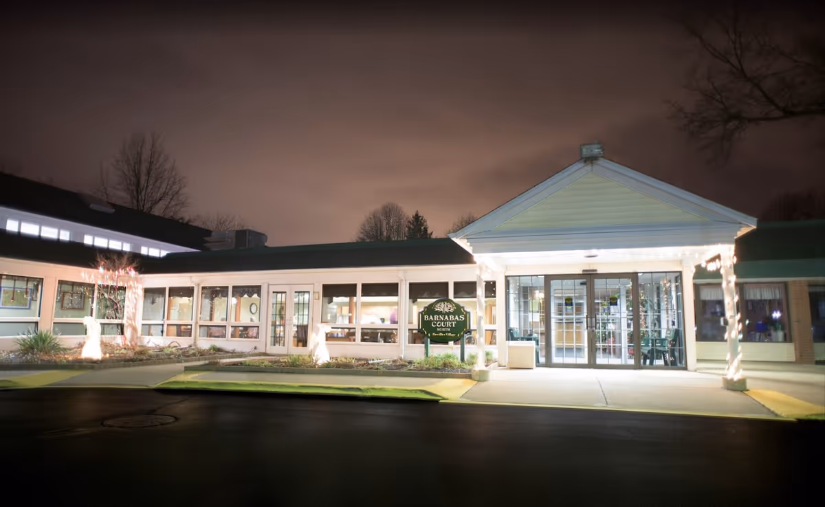 Night view of the entrance to Barnabas Court North at Brevillier Village, a single-story building with large windows and glass doors, decorated with string lights around the entrance pillars and illuminated landscaping.