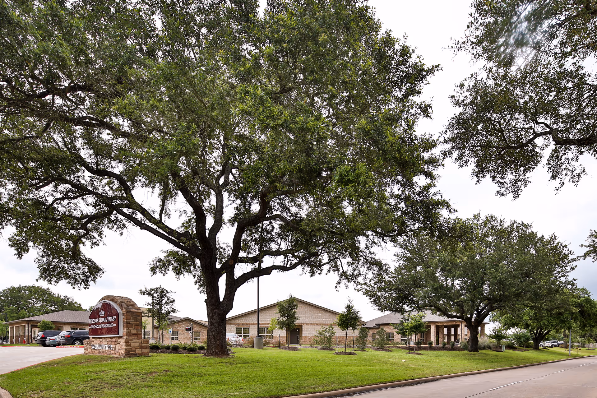 Single-story brick healthcare building with a stone entrance sign, large oak trees, and a grassy front lawn.