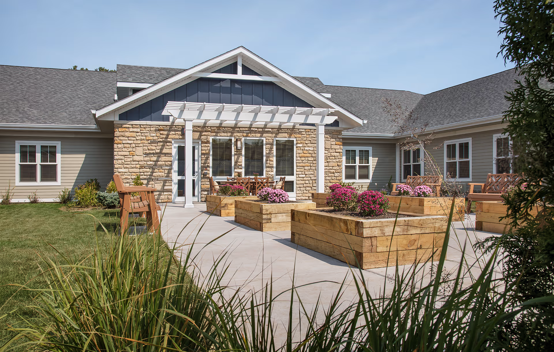 Outdoor patio area of a senior living facility with wooden raised garden beds filled with blooming flowers, wooden benches, and a stone and siding building with a pergola over the entrance.