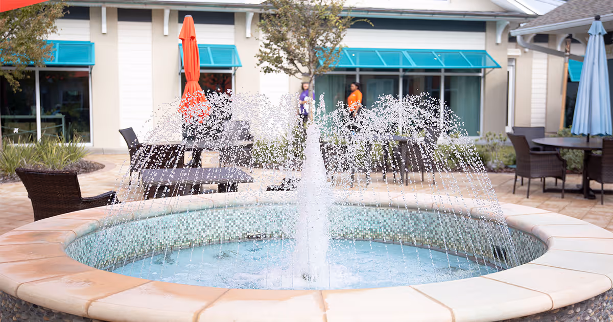 Round decorative fountain in a sunny courtyard with patio tables, umbrellas, and building windows in the background.
