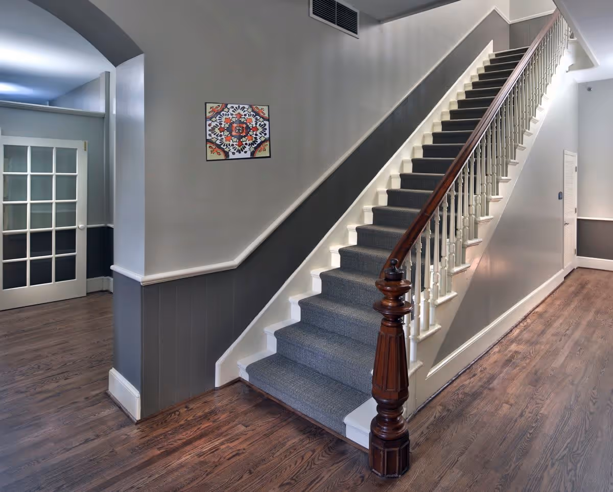 Carpeted staircase with a polished wooden banister in a clean interior hallway with gray walls, hardwood floors, and a glass-paneled door.