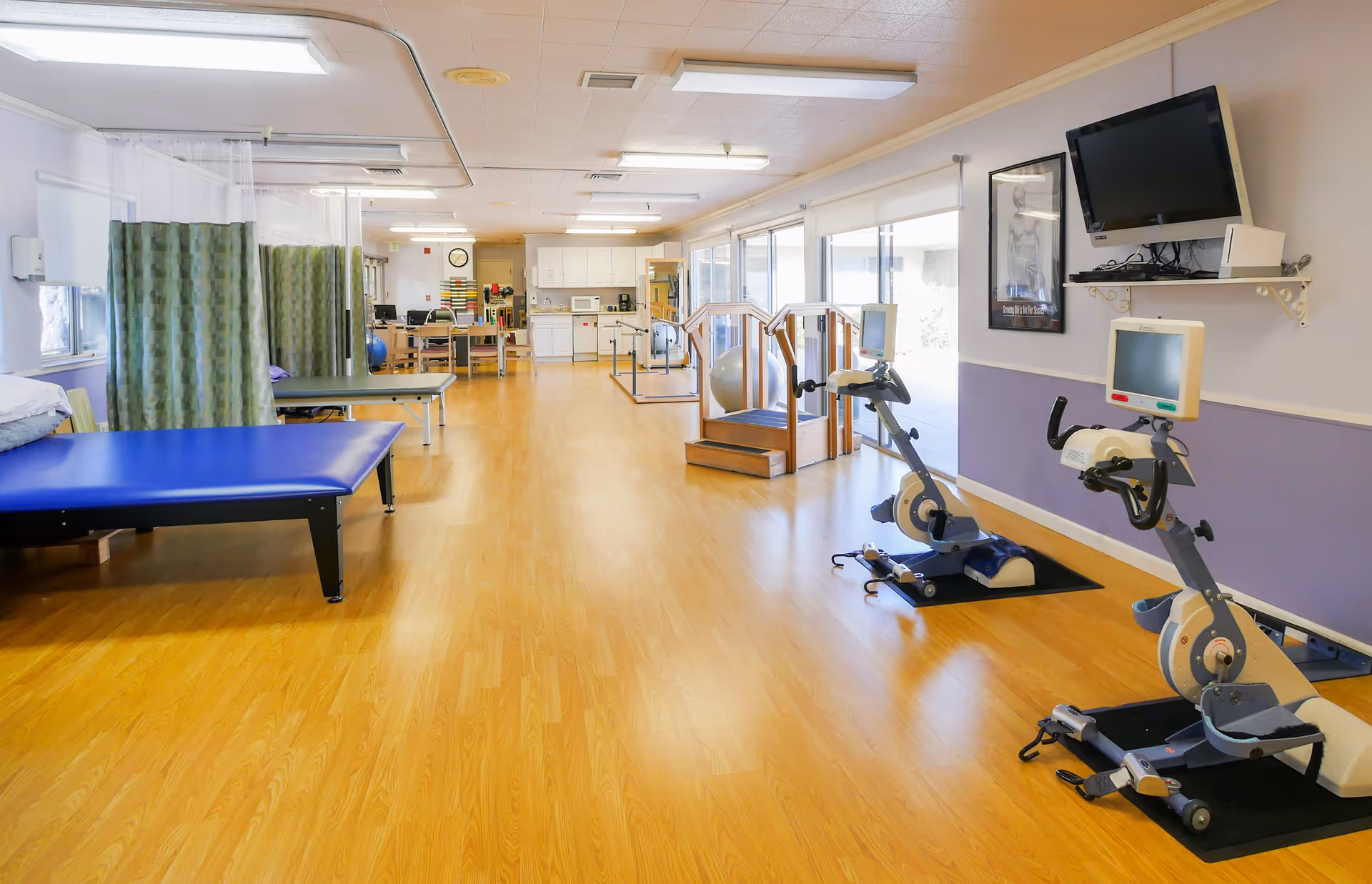 A spacious rehabilitation room with wooden flooring, featuring two stationary exercise bikes with monitors, parallel bars for walking exercises, a blue padded therapy table, privacy curtains, and a kitchenette area in the background.