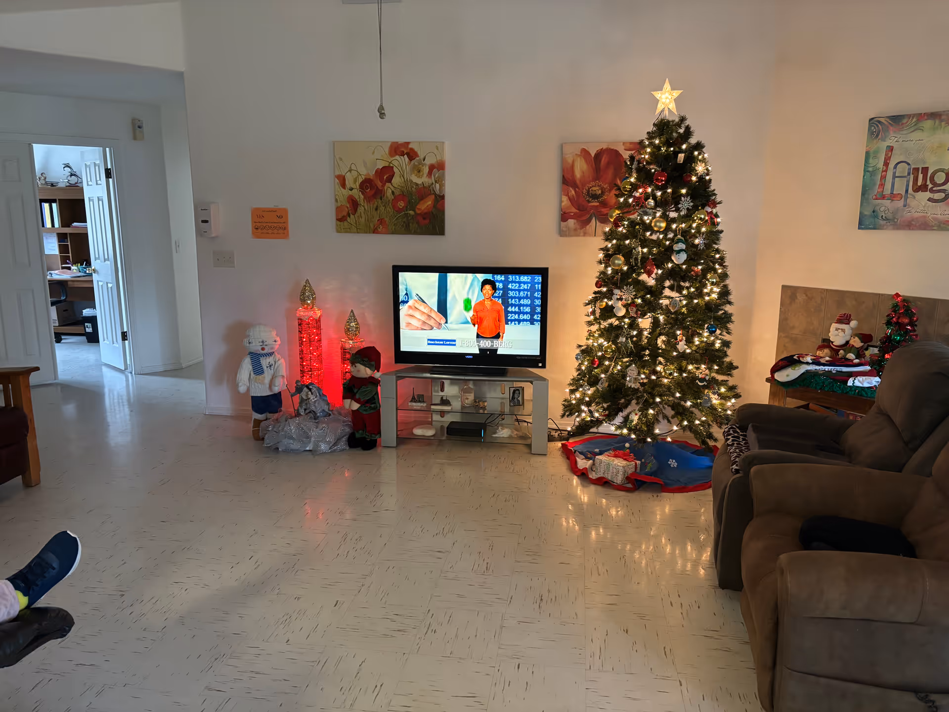 A cozy living room decorated for Christmas with a lit Christmas tree adorned with ornaments and a star on top. There is a flat-screen TV on a stand showing a person in an orange shirt, two festive figurines next to the TV, and a brown recliner chair on the right side. The walls have floral paintings and a colorful 'Laugh' sign. The floor is tiled, and an open doorway leads to another room with a desk and chair.