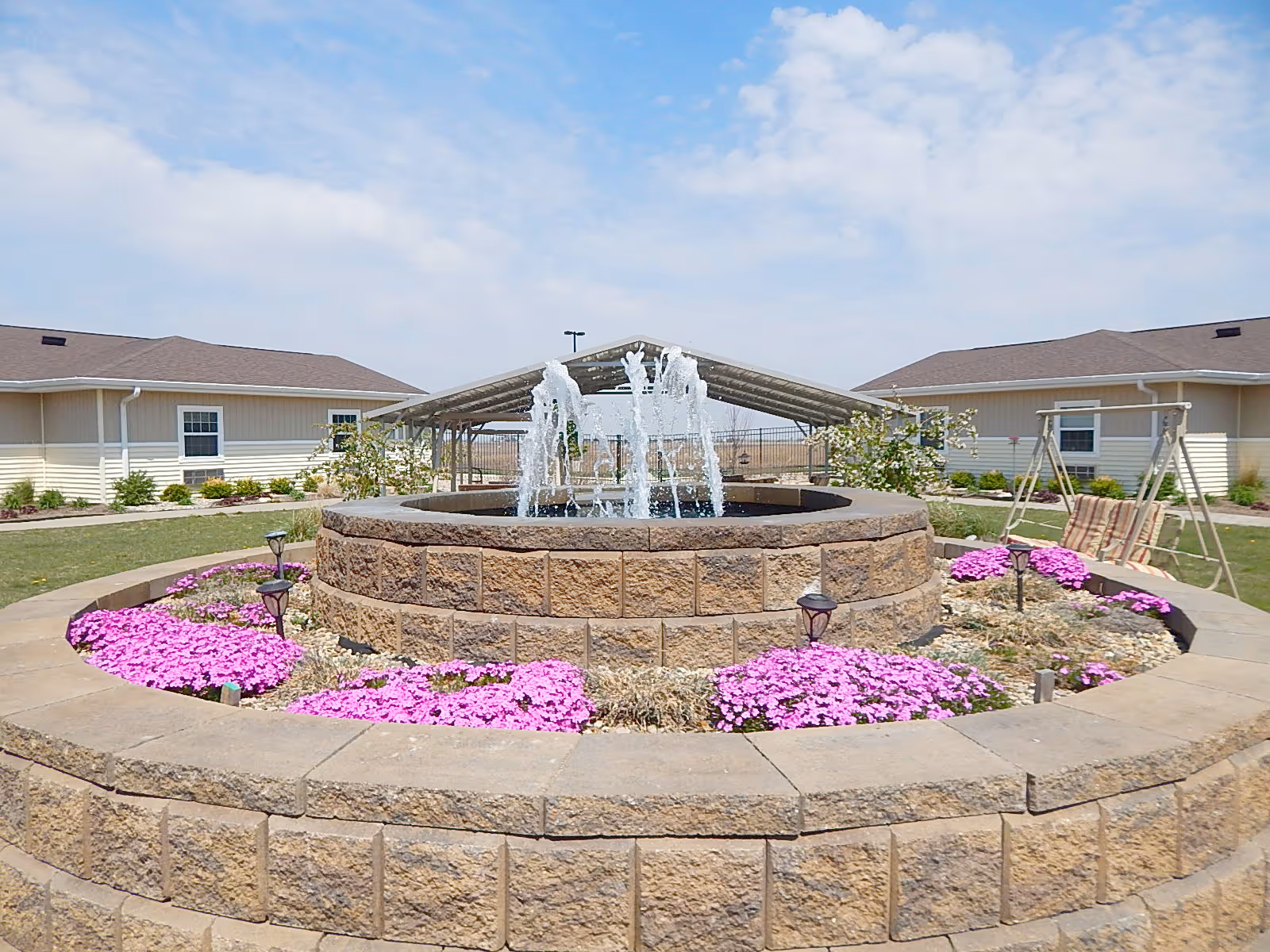Outdoor courtyard area with a circular stone water fountain in the center surrounded by pink flowers and landscaping. In the background, there are two single-story beige buildings with brown roofs, a covered seating area, and a swing bench. The sky is partly cloudy.