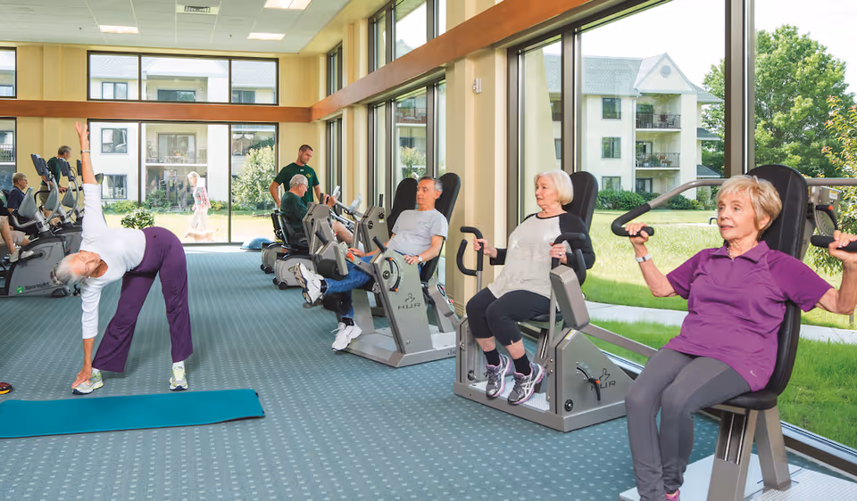 A group of older adults exercising on fitness machines and stretching in a bright, windowed exercise room overlooking the facility grounds.