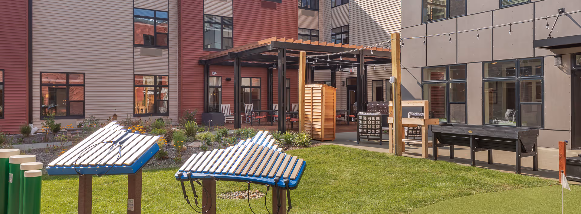 Outdoor courtyard area of a senior living facility with green grass, musical instruments including outdoor xylophones, a covered patio with seating, and a modern building facade with large windows in the background.