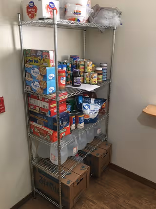 Metal shelving unit in a corner stocked with canned goods, boxed foods, gallon water jugs and paper products.