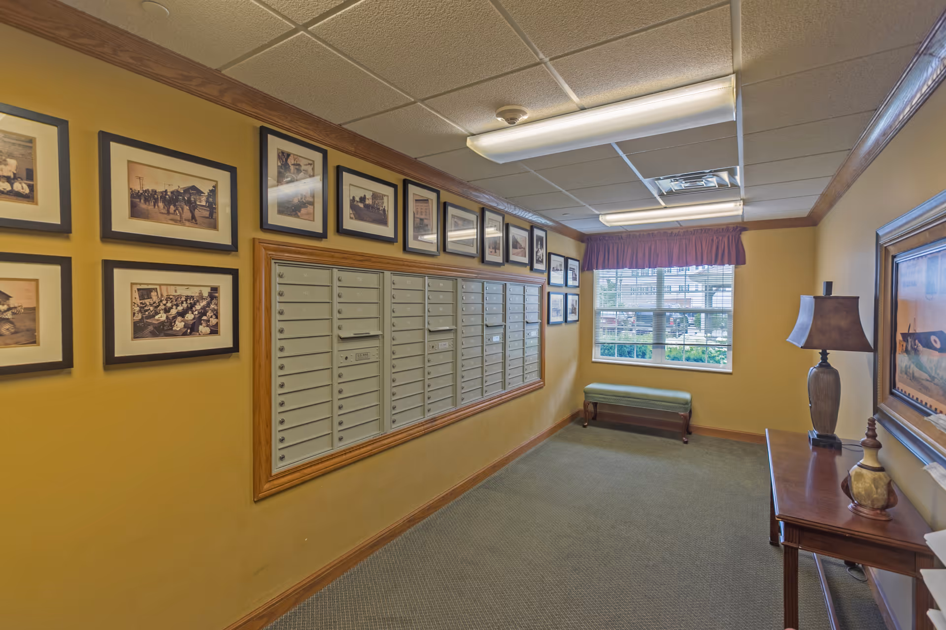 Interior hallway with a wall of mailboxes, framed vintage photographs above the mailboxes, a window with a purple valance, a green bench beneath the window, and a wooden table with a lamp and decorative items on the right side.