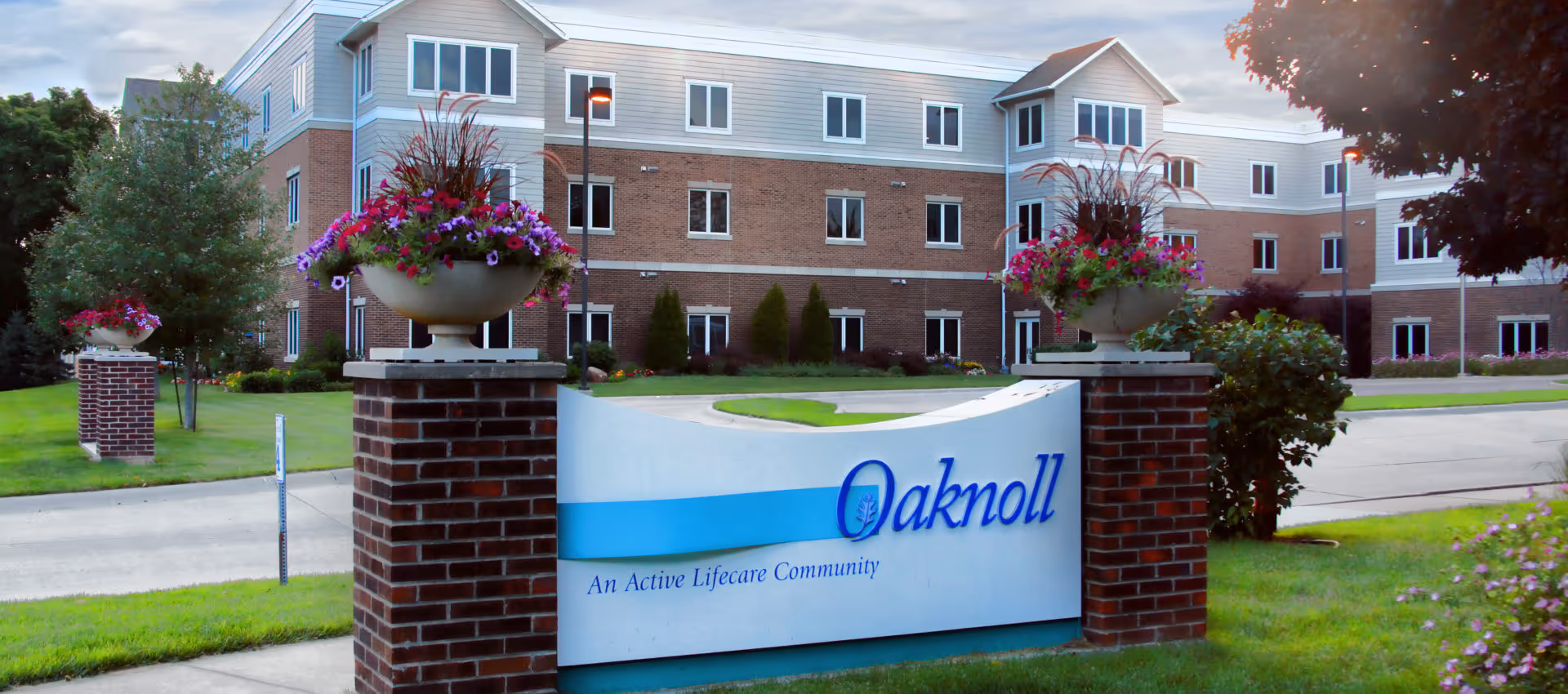 Entrance sign reading "Oaknoll — An Active Lifecare Community" in front of a brick multi-story retirement residence with decorative flower planters.