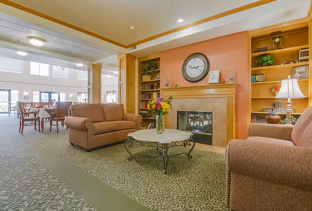 A cozy living room area with two brown upholstered sofas facing a fireplace with a clock above it. There is a round coffee table with a vase of colorful flowers in the center. Built-in wooden bookshelves flank the fireplace, holding books and decorative items. In the background, there is a dining area with tables and chairs near large windows letting in natural light.