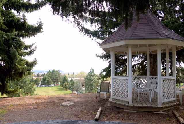 White wooden gazebo with chairs overlooking a grassy outdoor area and trees.