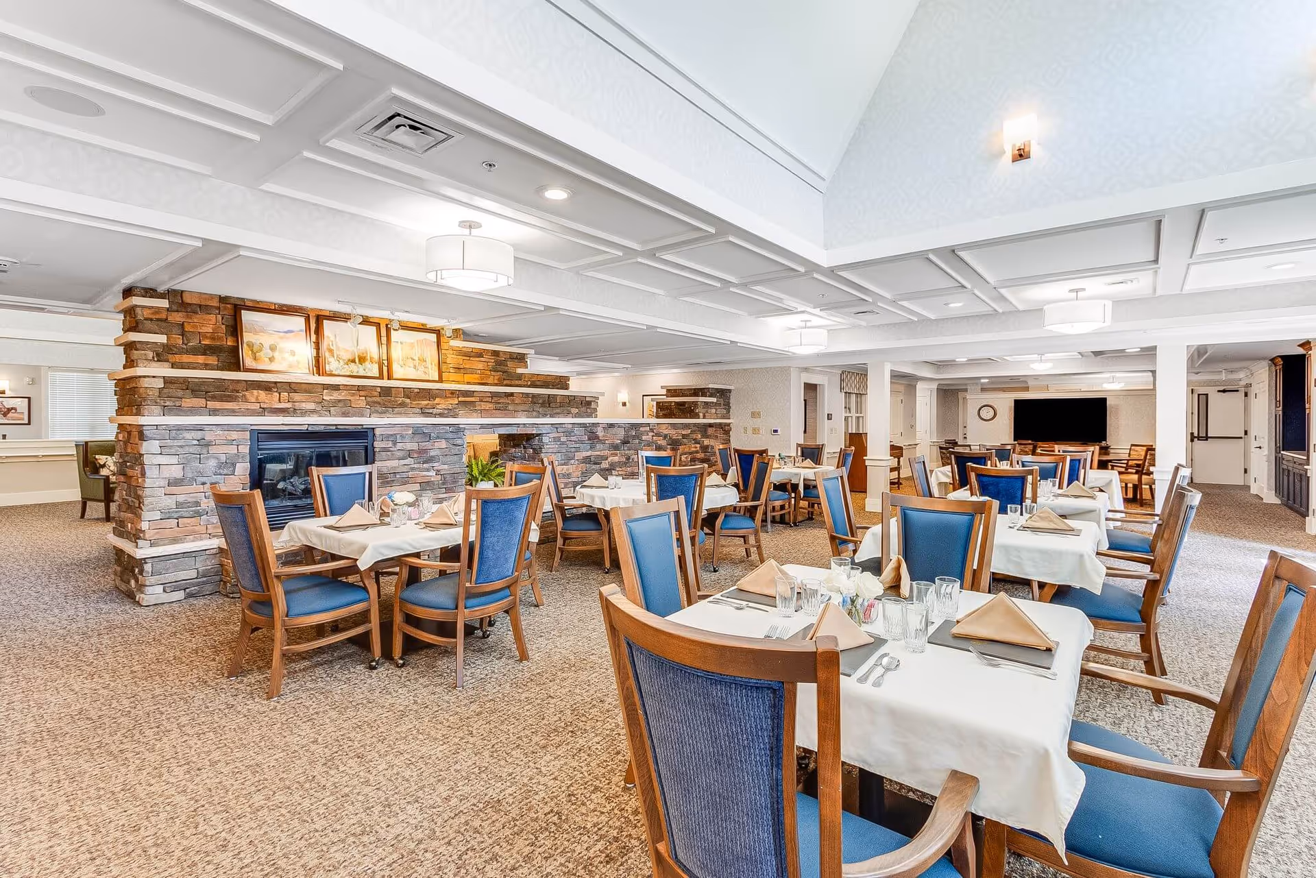 A spacious dining room in a senior living facility with multiple tables covered in white tablecloths, each set with beige napkins, glasses, and silverware. The room features blue cushioned wooden chairs, a stone fireplace with framed artwork above it, and a carpeted floor. The ceiling has recessed lighting and decorative panels.